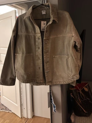 Beige jeansjacka från lagar 157 - Stilren beige jeansjacka från Weekday med klassisk krage, stora framfickor och knappar framtill. Jackan har en rak passform och är tillverkad i bomull, perfekt för dig som gillar en enkel men trendig look.