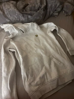 Ljusgrå hoodie från Lyle & Scott - Ljusgrå hoodie med klassisk huva och magficka från Lyle & Scott. På bröstet sitter den ikoniska gula örnloggan. Hoodien är tillverkad i mjukt bomullsmaterial och har ribbade muddar vid ärmslut och nederkant. Perfekt för en avslappnad och stilren look.
