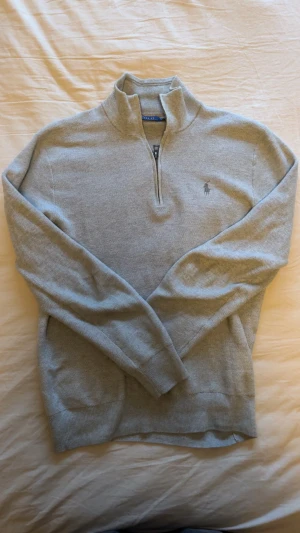 Grå halvzip tröja från Polo Ralph Lauren - Snygg ljusgrå stickad tröja från Polo Ralph Lauren med halv dragkedja och hög krage. Tröjan har det klassiska broderade logotypen på bröstet och ribbade muddar vid ärmslut och nederkant. Perfekt för lager på lager och stilren look.