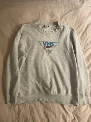 Grå sweatshirt med VHS Vision-tryck - Säljer en ljusgrå sweatshirt med retroinspirerat VHS Vision-tryck i blått och rosa på bröstet. Tröjan har rund halsringning, långa ärmar och mjukt material. Perfekt för dig som gillar vintage vibes och streetwear.