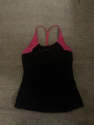Svart och rosa träningslinne - Säljer ett svart och rosa träningslinne från SOC med smala axelband och brottarrygg. Linnet är gjort i ett stretchigt funktionsmaterial som andas, perfekt för gymmet eller löpning. Snygg kontrast mellan svart tyg och rosa detaljer vid axlar och kanter.