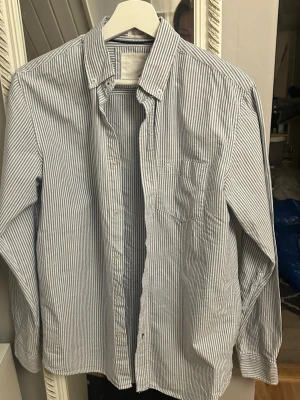 Randig skjorta med button-down krage - Säljer en klassisk skjorta med smala blå och vita ränder, button-down krage och bröstficka. Skjortan har långa ärmar och stängs med vita knappar framtill. Perfekt för en clean och stilren look.