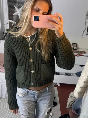 Mörkgrön stickad kofta med knappar - Mörkgrön stickad kofta i mycket fint skick. Grov ribbad struktur som ger en exklusiv och trendig look. Två stora fickor framtill i avvikande material som blir en snygg detalj. Knäppning med knappar hela vägen framtill. Perfekt till höst, vinter och lager-på-lager. 