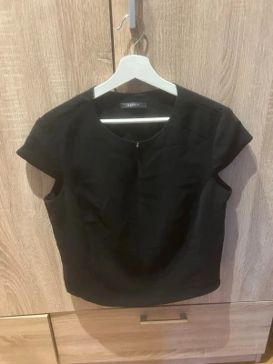 Svart kortärmad topp från Esprit - Stilren svart topp från Esprit med rund halsringning och korta ärmar. Modellen är croppad och har en enkel, klassisk design som passar till mycket. Perfekt att matcha med både jeans och kjol för en trendig look.