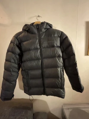 Svart sail racing dunjacka med huva - Säljer en svart sail racing dunjacka med huva och dragkedja framtill. Jackan har en puffig design med breda kanaler och två sidofickor. Perfekt för kalla vinterdagar och riktigt skön att bära. Passar dig som gillar en stilren och enkel look.