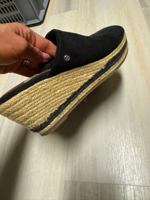 Svarta platåtofflor med jutesula - Säljer ett par svarta tofflor med hög platåsula i flätad jute och svart detaljrand. Ovandelen är i svart tyg och har en liten metallknapp på sidan. Perfekta för dig som vill ha en trendig och bekväm look.