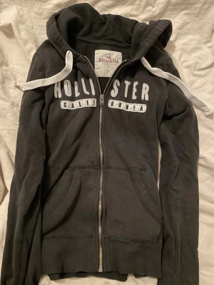 Svart zip-hoodie från Hollister - Mysig svart hoodie från Hollister med dragkedja, stora fickor och vit broderad text 'Hollister California' på bröstet. Hoodien har vit dragsko i huvan och är gjord i mjukt material, perfekt för chill dagar. Klassisk streetwear-vibe!