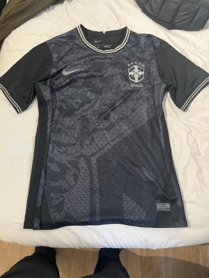 Svart Brasilien Nike fotbollströja - Snygg svart fotbollströja från Nike med Brasilien-logga på bröstet och diskret mönster över hela framsidan. Tröjan har rund hals, korta ärmar och vita detaljer vid krage och ärmslut. Perfekt för träning eller match.