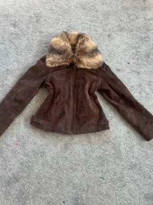 Brun skinnjacka med fuskpäls - Snygg brun skinnjacka från Bik Bok med dragkedja framtill och två fickor med dragkedja. Jackan har en stor, fluffig fuskpäls-krage i beige och bruna toner som ger en lyxig känsla. Figursydd modell med justerbara spännen vid midjan. Perfekt för dig som vill sticka ut.