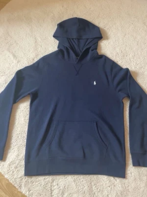 Blå fleece hoodie från Polo Ralph Lauren - Mörkblå hoodie från Polo Ralph Lauren med klassisk huva och känguruficka framtill. Liten vit broderad logga på bröstet. Tillverkad i mjukt material som känns skönt mot huden. Perfekt för en avslappnad och stilren look.