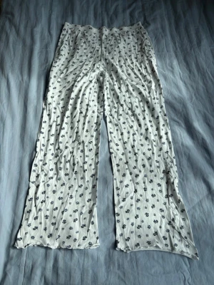 Vita pyjamasbyxor med blommönster - Säljer ett par vita pyjamasbyxor med små blå blommor över hela tyget. Byxorna har en lös och bekväm passform med raka ben, perfekta för att chilla hemma. Materialet känns mjukt och svalt mot huden, vilket gör dem sköna att ha på sig under natten.