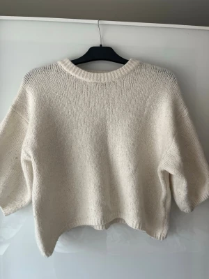 Ginatricot - Säljer en krämvit stickad tröja med korta ärmar och rund hals. Tröjan har en avslappnad, boxig passform och är tillverkad i ett mjukt stickat material. Perfekt för lager på lager och enkel att styla till olika outfits.