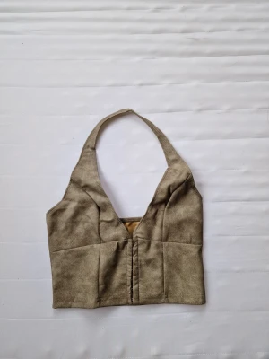 Beige halterneck linnetopp - Trendig beige halterneck linnetopp med djup v-ringning och croppad passform. Toppen har en enkel design utan mönster och är tillverkad i ett mjukt tyg som känns bekvämt mot huden. Perfekt att styla med höga jeans eller kjol.