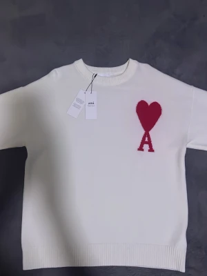 Vit stickad tröja  - Vit stickad tröja  med röd hjärt- och A-detalj på bröstet. Tröjan har rund halsringning, ribbade muddar och en avslappnad passform. Perfekt för dig som gillar stilrena plagg med en twist. Fråga gärna om fler bilder 