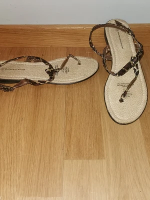 Leopardmönstrade sandaler med remmar - Snygga sandaler med tunna remmar i leopardmönster och beige sula. Remmarna är justerbara och ger en trendig look. Perfekta för sommaren och lätta att matcha med olika outfits. Sulan är platt och bekväm för vardagsbruk.