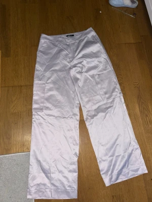Lavdender vida linnebyxor med fickor - Säljer ett par ljuslila cargobyxor med raka ben och stora sidofickor. Byxorna har en avslappnad passform och är tillverkade i ett glansigt, mjukt material som ger en cool look. Perfekta för dig som gillar streetwear och vill ha något bekvämt men ändå stilrent.