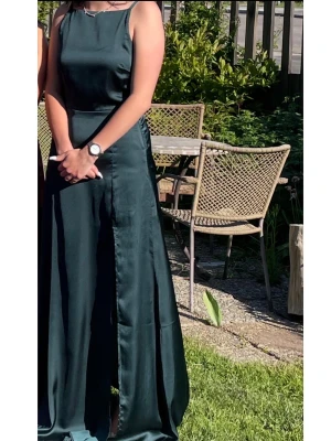 Mörkgrön långklänning med slits i storlek 36 - Elegant mörkgrön långklänning i ett glansigt material med smala axelband och slits framtill. Klänningen har en enkel, stilren design med rak halsringning och figurnära passform upptill. Djup i ryggen med spetskant (bild 3). Är använd vid ett tillfälle och är i fint skick utan några skavanker. Perfekt för speciella tillfällen.