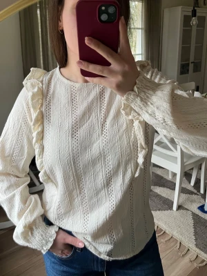 Blus med volang  - Söt vit/beige blus med långa ärmar och dekorativa volanger längs axlarna samt ett hålmönster. Passar även M skulle jag säga.