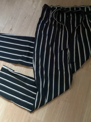 randiga kostymbyxor med knyt - Randiga kostymbyxor med vita vertikala ränder och knytband i midjan. Byxorna har raka ben och en avslappnad passform, perfekta för en stilren look. Materialet känns mjukt och följsamt.