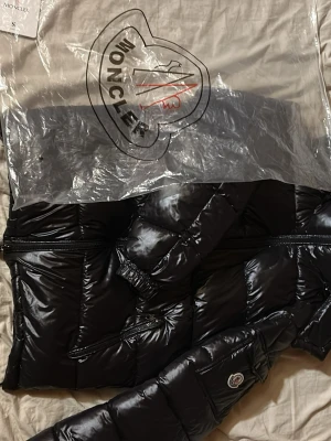 Svart moncler maya - Säljer en svart, glansig dunjacka från Moncler med klassisk quiltad design och dragkedja framtill. Jackan har hög krage och Moncler-logga på ärmen. Perfekt för kalla vinterdagar och riktigt snygg streetstil.