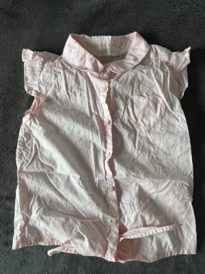 Ljusrosa skjorta med hjärtficka H&M - Söt ljusrosa skjorta från H&M med korta ärmar och volangdetaljer. Skjortan har klassisk krage, knäppning framtill och en hjärtformad bröstficka. Perfekt för en lekfull och trendig look.