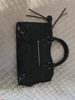 Svart handväska med flätat handtag - Snygg svart handväska i skinnimitation med flätat handtag och silverfärgade metalldetaljer. Väskan har en framficka med dragkedja och dekorativa nitar samt långa läderband på dragkedjorna. Perfekt för dig som gillar en edgy och stilren look. Helt oanvänd!!!!