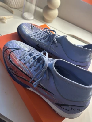 Nike Mercurial fotbollsskor/inomhusskor - Säljer ett par Nike Mercurial fotbollsskor i ljusblått/lila med svarta och röda detaljer. Skorna har hög krage för extra stöd runt ankeln och är tillverkade i ett syntetmaterial med textur för bättre bollkontroll. Snörning framtill. Jag tycker dom funkar jätte bra men nu har jag större storlek så dom passade tyvärr intr längre. Bekväma!  Skriv om ni har några frågor❤️
