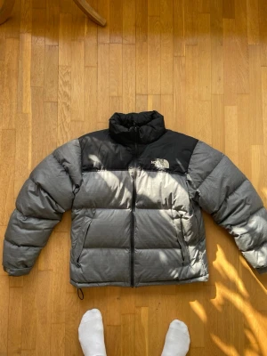 Grå och svart pufferjacka North Face - Säljer en grå och svart pufferjacka från The North Face med hög krage och dragkedja. Jackan har två sidofickor med dragkedja och logotyp på bröstet. Perfekt för kalla vinterdagar, tillverkad i slitstarkt material och fylld för extra värme. Nypriset är runt 4000kr.
