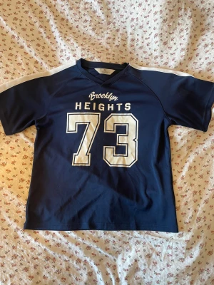 Mörkblå  t-shirt - Säljer en mörkblå t-shirt med texten ”Brooklyn Heightd 73” på på framsidan. T-shirten har korta ärmar och en vit detalj längs axlarna. Perfekt för dig som gillar sportig stil och vill sticka ut! Är i storlek 134/140 men den passar mig bra som är en xs-s!! 💙