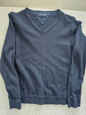 Mörkblå stickad tröja Tommy Hilfiger - Mörkblå stickad tröja från Tommy Hilfiger med V-ringning och diskret logga på ärmen. Tröjan har långa ärmar och ribbade muddar vid ärmslut och nederkant. Perfekt för lager-på-lager och enkel att matcha med jeans eller chinos.