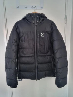 Svart pufferjacka från Haglöfs - Säljer en svart pufferjacka från Haglöfs med huva och dragkedja framtill. Jackan har en bröstficka med Haglöfs-logga och justerbar dragsko till hu', samt quiltad design för extra värme. Perfekt för kalla vinterdagar och riktigt skön att bära. Den är lite sliten med små skador som knappt märks. Pris kan diskuteras. Fraktas inom 24 timmar.