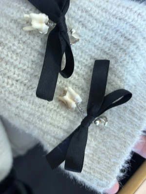 Örhängen med svarta rosetter och tänder - Unika örhängen med svarta sidenrosetter och hängande tänder som dekoration. Fästet är i silverfärgad metall och designen är edgy och alternativ. Perfekt för dig som vill sticka ut med accessoarer som verkligen syns.