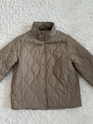 Brun quiltad pufferjacka - Snygg brun pufferjacka med vågigt quiltat mönster och hög krage. Jackan har tryckknappar framtill och raka ärmar. Köpt på Åhléns och är helt ny!🫶🏻
