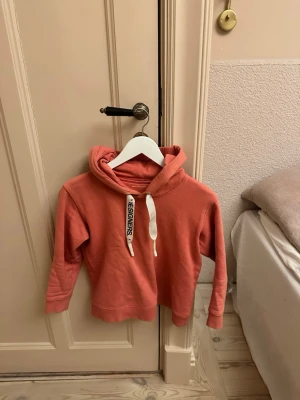 Korall hoodie från Designers Remix - Stilren hoodie i korallrosa från Designers Remix med vita dragsnören där det står 'DESIGNERS' i svart text. Klassisk huva och ribbade muddar vid ärmar och nederkant. Perfekt för en chill och trendig stil.