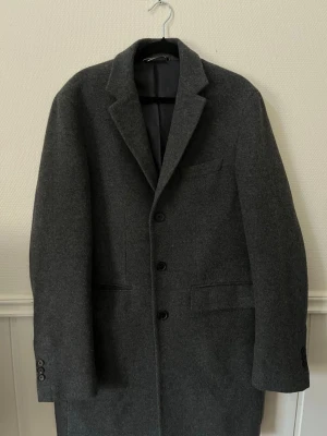 Mörkgrå gant ullrock med klassisk krage - Nypris 4000kr. Stilren mörkgrå ullrock från gant med tre knappar framtill, klassisk krage och två fickor med lock. Rocken har en rak passform och slits bak. Perfekt för kyliga dagar och ger en snygg, tidlös look.