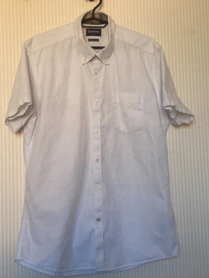 Vit kortärmad skjorta från Barbour - Klassisk vit skjorta från Barbour med korta ärmar och bröstficka. Skjortan har knappar hela vägen fram och en stilren button-down krage. Perfekt till sommaren och enkel att matcha med olika outfits.