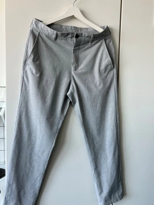 Ljusgrå chinos med diskret mönster - Snygga ljusgrå chinos med diskret rutmönster och raka ben. Byxorna har bälteshällor, knapp och dragkedja i midjan samt två sidofickor. Materialet känns mjukt och bekvämt. Nypris: 429kr 