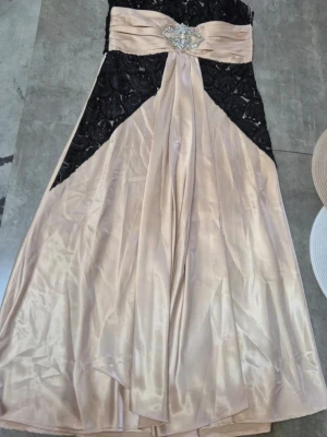 Långklänning i svart och champagne - Elegant långklänning i svart spets och champagnefärgad satin. Klänningen har axelbandslös design, markerad midja med draperad detalj och glittrigt smycke framtill. Perfekt för speciella tillfällen där du vill sticka ut.