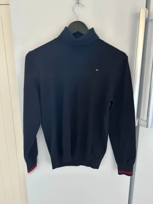 Svart polotopp från Tommy Hilfiger - Klassisk svart stickad polotopp från Tommy Hilfiger med diskret logga på bröstet. Tröjan har långa ärmar och ribbade muddar med röda och marinblå detaljer. Perfekt till höst och vinter för en stilren look.