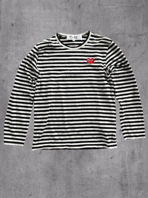 Randig långärmad topp från Comme  des Garçons - Säljer en svartvit randig långärmad topp från Comme des Garçons PLAY med det ikoniska röda hjärtat broderat på bröstet. Tillverkad i mjuk bomull och har en klassisk passform. Perfekt för dig som gillar streetstyle och vill ha något tidlöst. Liten i storleken mer XS