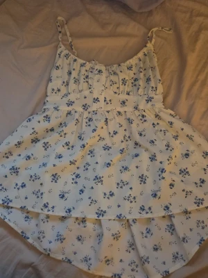 Blommig vit och blå topp - Söt topp med blå blommönster på vit botten. Toppen har tunna axelband, knappar framtill och en fin knytning i ryggen. Perfekt för varma dagar och ger en romantisk vibe till din outfit.