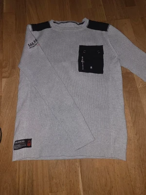 Ljusgrå stickad tröja Sail Racing - Ljusgrå stickad tröja från Sail Racing med svarta axelpartier och en svart bröstficka med knappdetaljer. Tröjan har ribbstickad struktur, rund hals och tryck på ena ärmen samt en patch nertill. Storlek M, storlekslappen är borta men det är M herr. Använd ett fåtal gånger.