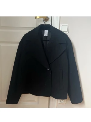 Svart kappa från C.W, storlek 34 - Stilren svart kappa från C.W i storlek 34. Kortare modell med bred krage och diskret knappstängning framtill. Kappan har raka ärmar och två fickor framtill. Perfekt för dig som gillar minimalistisk och klassisk stil.
