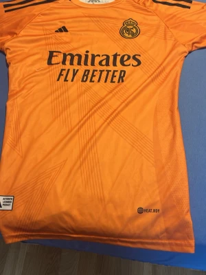 Orange Real Madrid målvaktströja - Säljer en orange Real Madrid målvaktströja från Adidas med svarta detaljer och klubbmärke på bröstet. Tröjan har 'Emirates Fly Better' tryckt på framsidan och klassiska Adidas-ränder på axlarna. Tillverkad i lätt och ventilerande material.