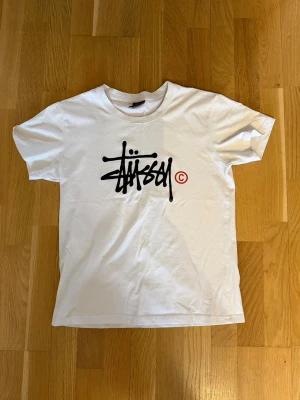 Vit Stüssy t-shirt med logga - Säljer en vit t-shirt från Stüssy med deras ikoniska svarta logga och röd copyright-symbol på bröstet. Klassisk passform med rund hals och korta ärmar. Tillverkad i mjuk bomull, perfekt för en avslappnad streetwear-look.