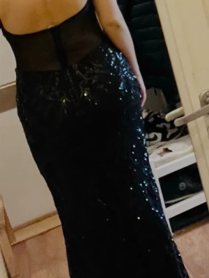 Svart glittrig långklänning med slits - Säljer en svart långklänning med glittriga paljetter, hög slits framtill och mesh-detaljer vid bysten och ryggen. Klänningen är ärmlös och har en elegant, figurnära passform som ger en snygg siluett. Perfekt för bal eller festliga tillfällen.