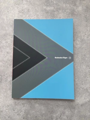 Onitsuka Tiger broschyr - Snygg broschyr från Onitsuka Tiger med grafisk design i blått, svart, grönt och rött. Omslaget har zigzag-mönster och logotypen syns tydligt både fram och bak. Perfekt för dig som gillar sneakers, streetwear eller samlar på märkesrelaterade trycksaker.