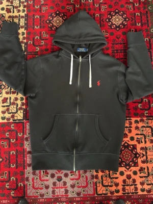 Svart hoodie från Polo Ralph Lauren - Svart hoodie med dragkedja från Polo Ralph Lauren. Klassisk design med vit dragsko i huvan och röd broderad logga på bröstet. Mjuk insida och två fickor framtill. Perfekt för en avslappnad och stilren look.