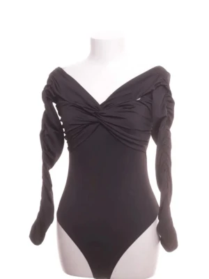 Body - Trendig svart body med långa ärmar och off shoulder-design. Framtill har bodyn en snygg twistad detalj över bysten som ger en cool look. Materialet är stretchigt och följsamt, perfekt för att skapa en smickrande siluett. Passar dig som gillar stilrena och moderna plagg.