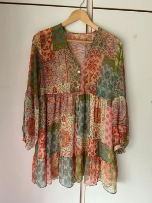 Patchwork klänning med v-ringning - Bohemisk kortklänning i patchworkmönster med inslag av grönt, rosa, orange och beige. Klänningen har v-ringning, knappar framtill och långa ärmar med resår vid ärmslut. Tillverkad i ett lätt och luftigt material, perfekt för varma dagar.